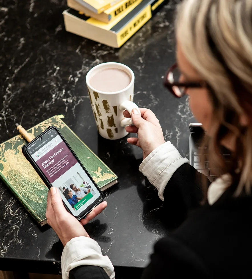 A person holding a coffee mug and smartphone, reading a website. A green notebook and stacked books are on the black marble table, evoking a cozy work scene.